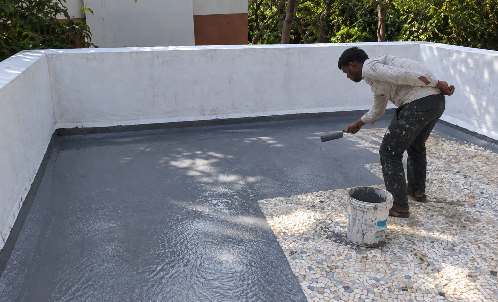 Worker roller-applying grey waterproof coating across a balcony floor up to the parapet
